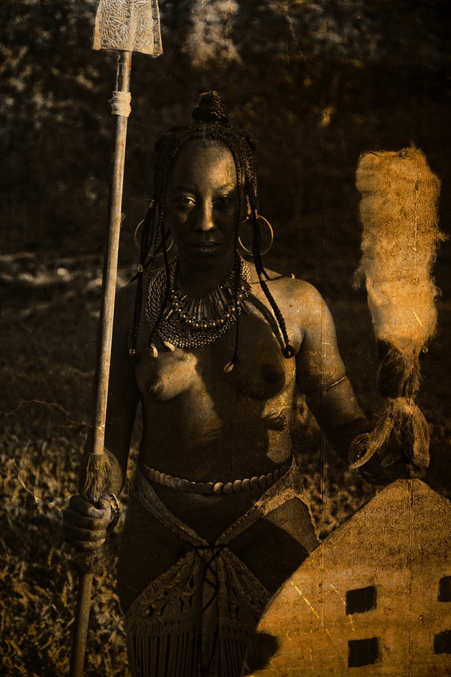 Photograph of J LeShae casted as Akofena in the Dynasty Photo Series by artist, Missy Burton. J is pictured in a field with a spear and shield, wearing several gold adornments on an otherwise topless body. Her hair is cornrowed and hangs to the shoulders. The photograph is a mixture of sepia tones