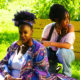 Picture of two Black African American women sitting outdoors. One woman is centered in the image, sitting in the grass wearing a large afro puff ponytail and a multicolored purple kaftan. The other woman is crouching behind her looking off screen to the left. She is wearing braids, and off the shoulder light pink shirt, and jeans. She has a camera hanging around her neck. They are surrounded by greenery.